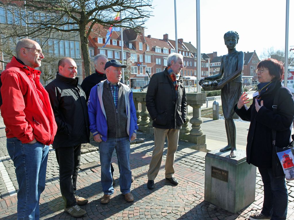 Eine Stadtführerein steht mit einer Gruppe vor der Bronzestatue Janjte Viss und erzählt etwas über die Stadtgeschichte