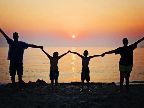 Zu sehen ist eine 4-köpfige Familie. Alle halten sich an den Händen und heben die Hände dabei in die Höhe. Sie stehen in einer Reihe und schauen in den Sonnenuntergang. Man erkennt nur dunkle Umrisse der Menschen.