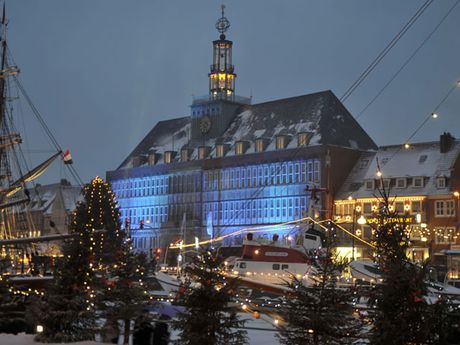 Beleuchtete Weihnachtsbaeume, dahinter der Seenotrettungskreuzer Georg Breusing und im Mittelpunkt das Ostfriesische Landesmuseum, leicht mit Schnee überzogen.