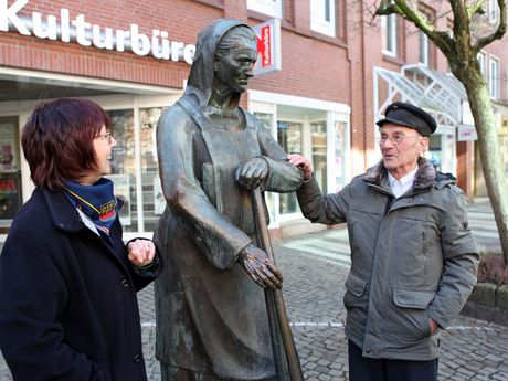 Eine Stadtführerin steht neben der Bronzefigur Peterke und erzählt einem interssierten Gast geschichtliches zu Emden